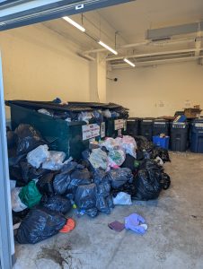 A commercial dumpster that is completely overloaded and blocked by an excessive amount of garbage bags